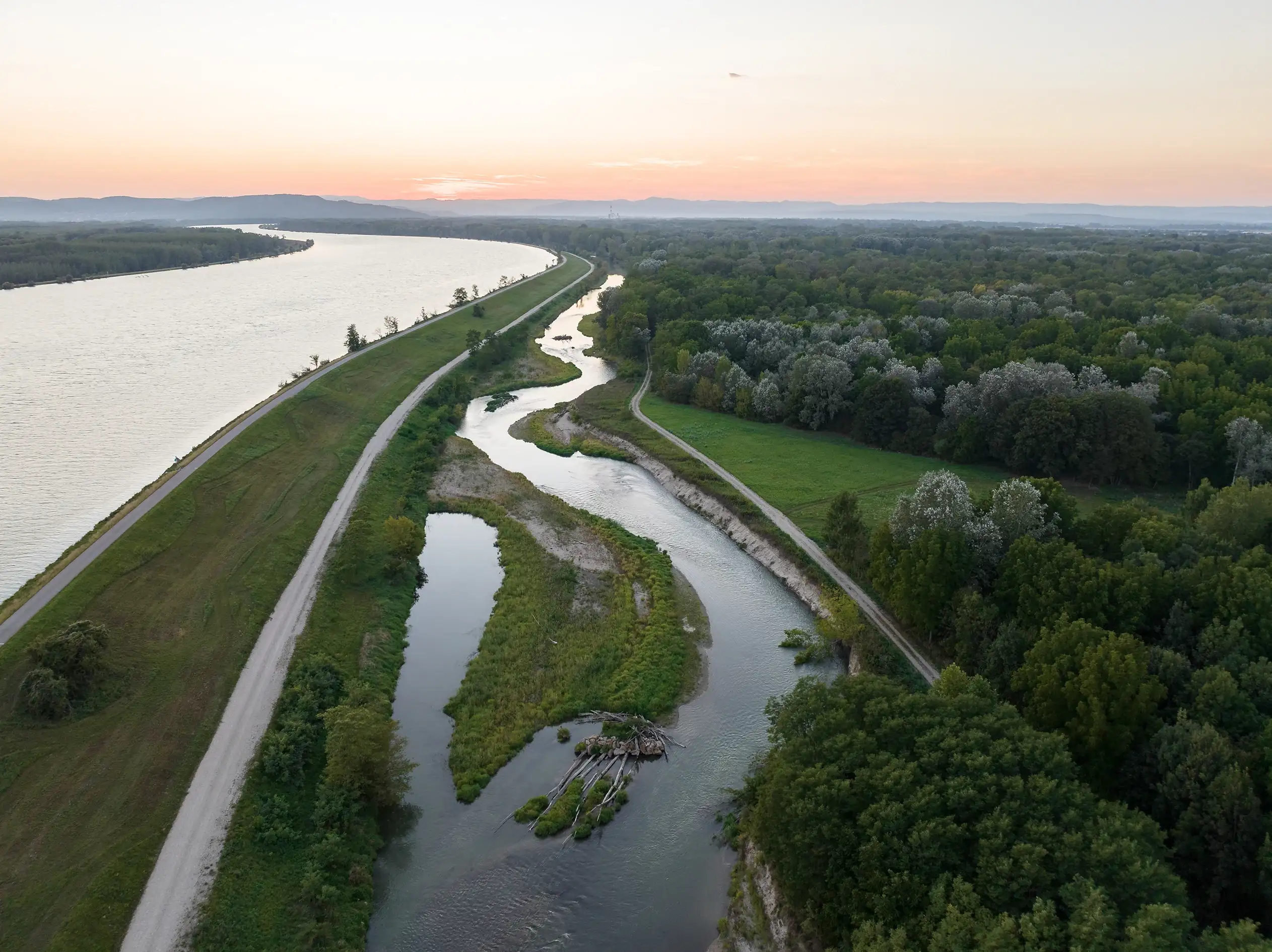 Blick auf die Fischwanderhilfe bei Wasserkraftwerkt Altenwörth bei Sonnenuntergang. Der Himmel ist in ein zartes Rosé getaucht.